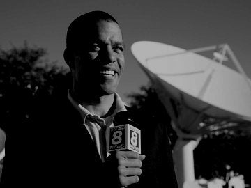Male reporter broadcasting live from outside a large broadcast studio, satellite dishes in background.